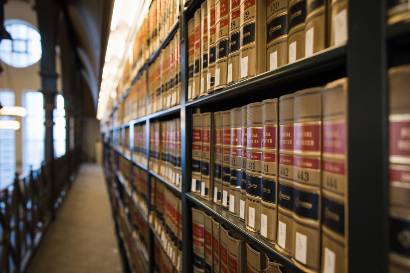 Rows of leather-bound law books line dark wooden shelves along a narrow library aisle, with arched windows beyond.