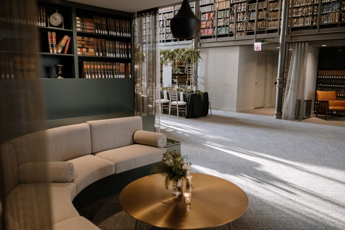 Curved cream sofa and round gold coffee table with flowers sit near tall bookshelves in a carpeted library lounge.
