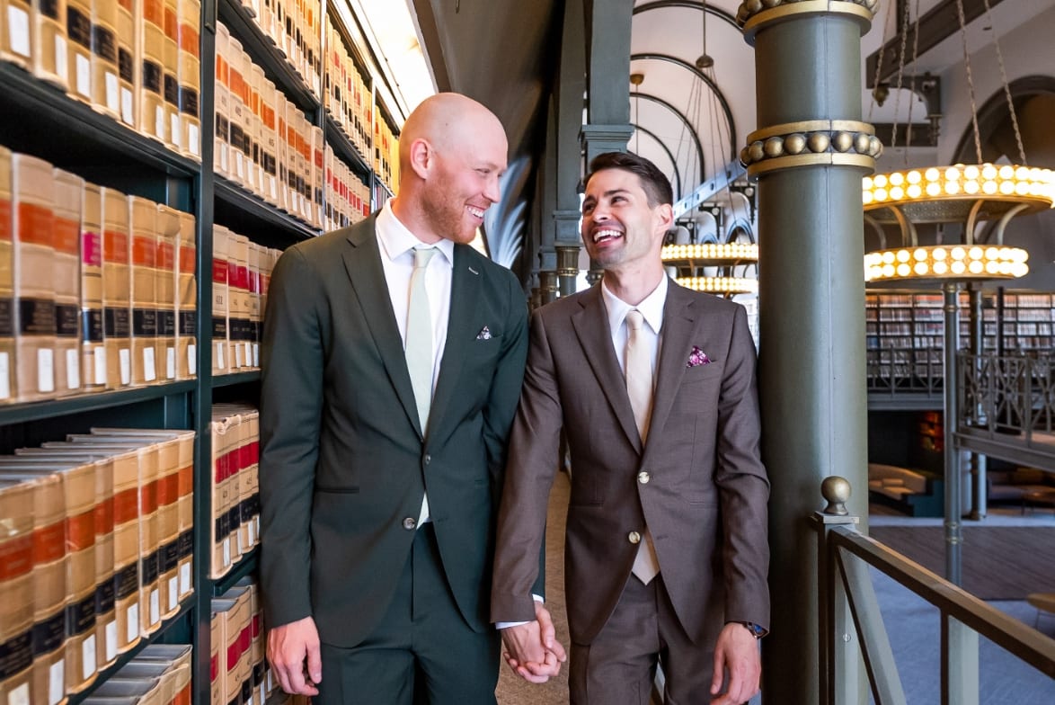 Two grooms holding hands beside the bookshelves