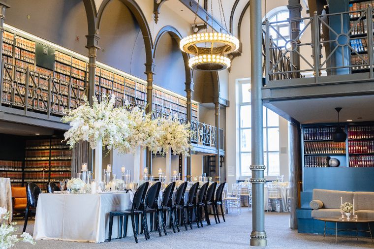 Long banquet table with white linens, tall white floral centerpieces and black chairs beneath arched library balconies.