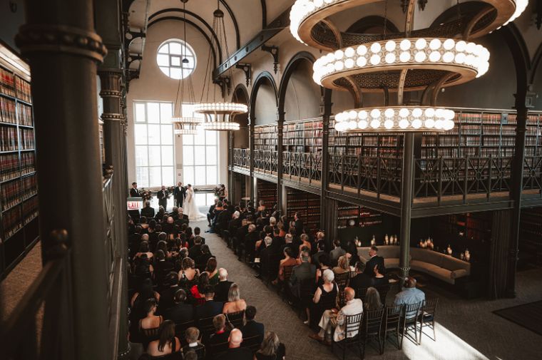 Wedding ceremony in a two-story library hall, with seated guests facing the couple beneath large ring chandeliers.
