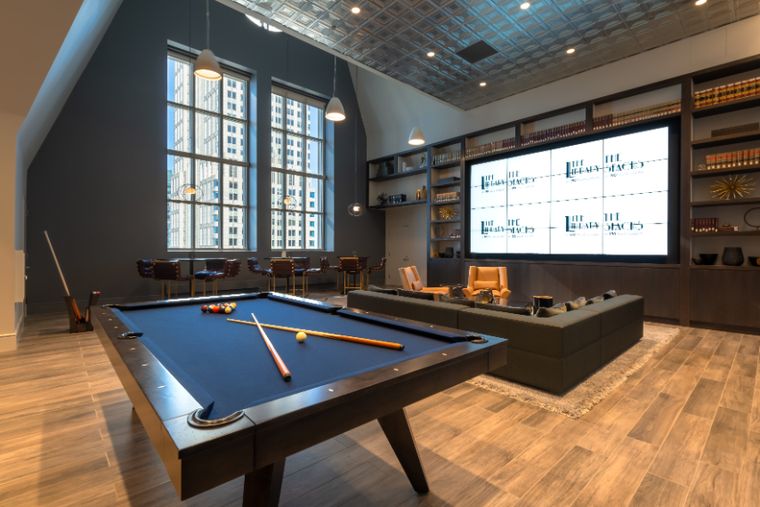 Blue-felt pool table in a lounge with tall arched windows, a video wall of screens, bookshelves, and leather sofas.