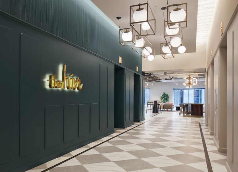 Corridor with a dark green paneled wall, gold "the hive" sign, cube pendant lights, and checkered tile floor.
