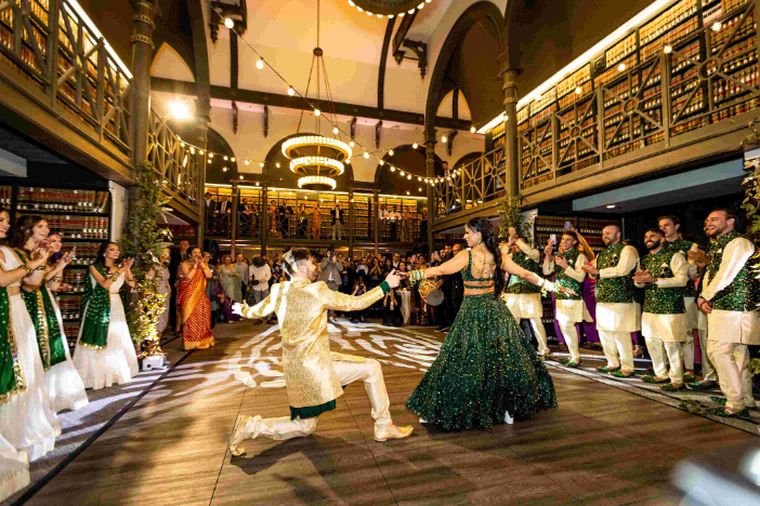 Couple in gold and green wedding attire dance on a wood floor as guests in matching outfits clap in a library hall.