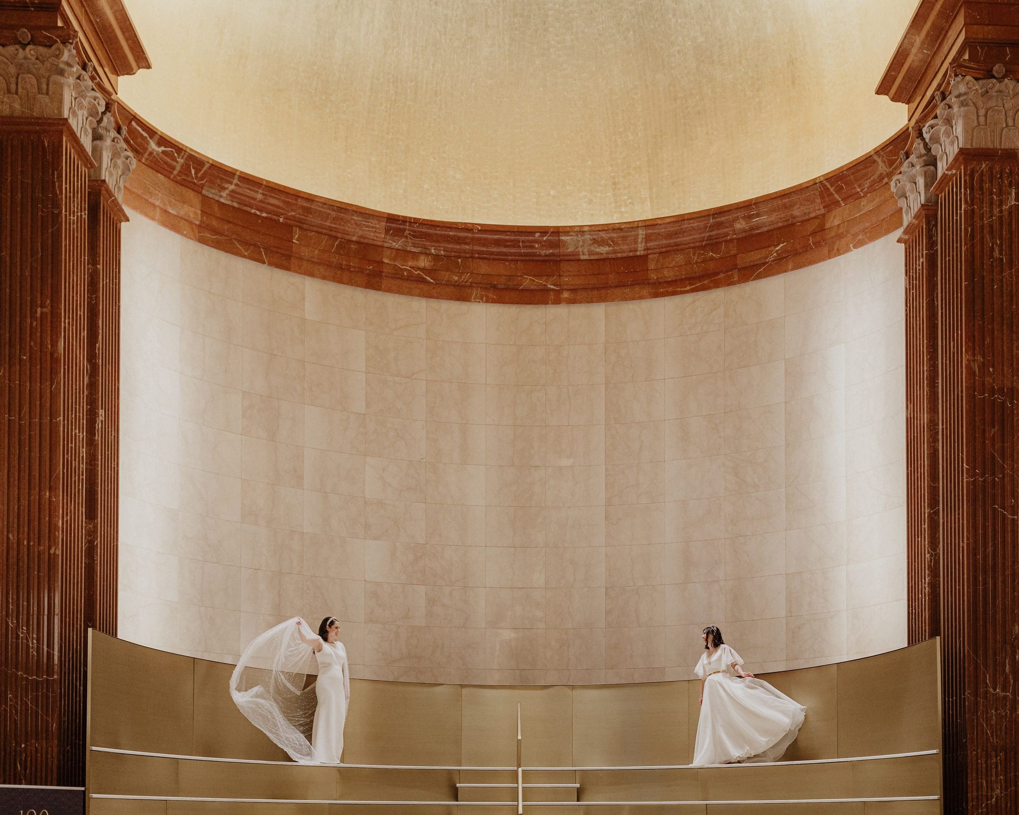Two women in white gowns stand at opposite ends of a balcony below a curved marble wall and gold domed ceiling.