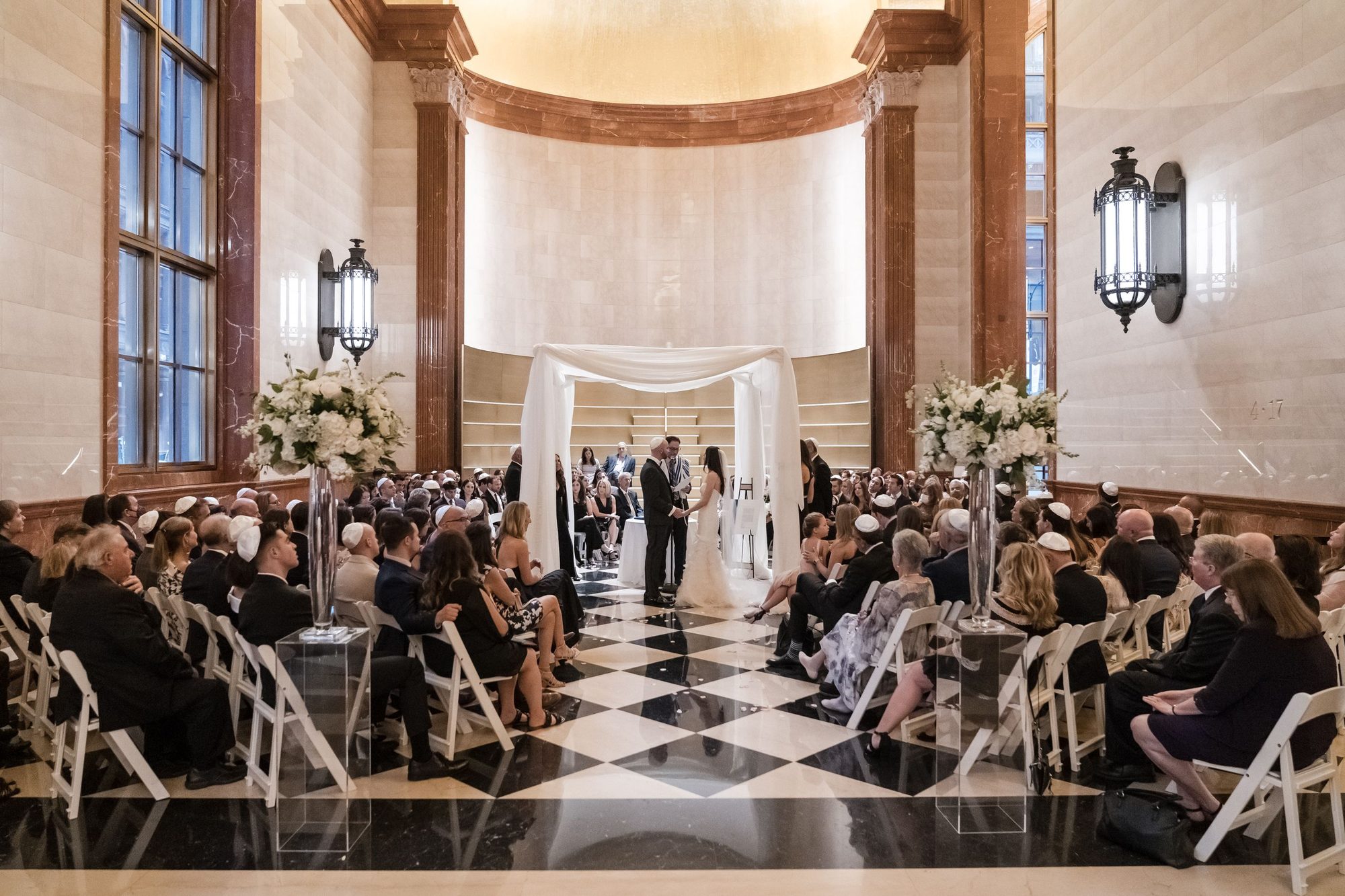 Bride and groom stand under a white chuppah as seated guests watch from white chairs in a marble hall with checkered floor.