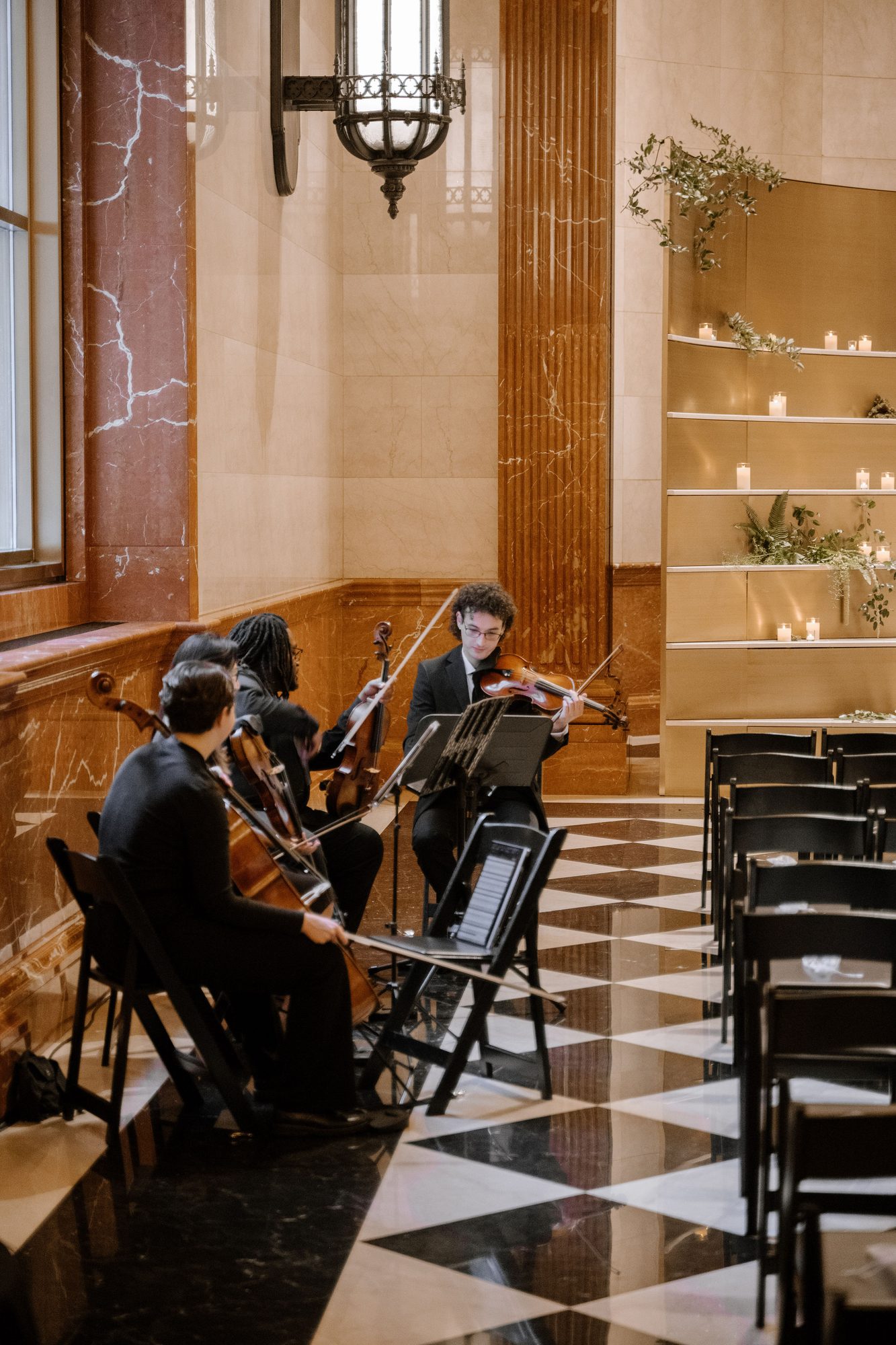 String quartet plays beside marble walls and candlelit shelves, with rows of chairs on a checkered floor.