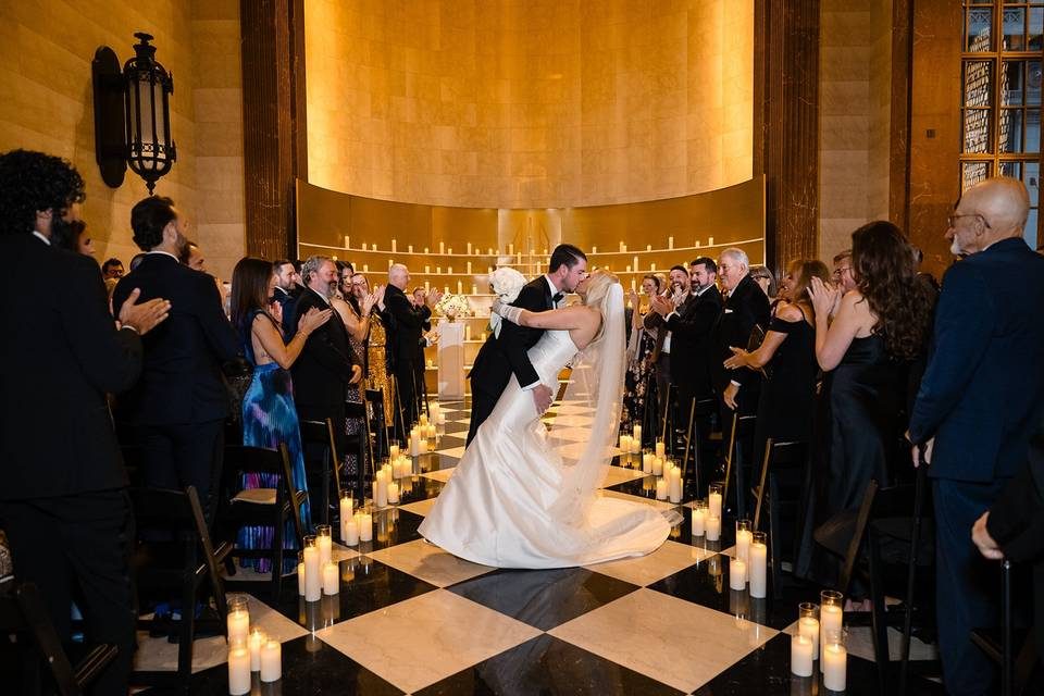 Bride and groom kiss on a checkered marble aisle lined with pillar candles as standing guests clap on both sides.