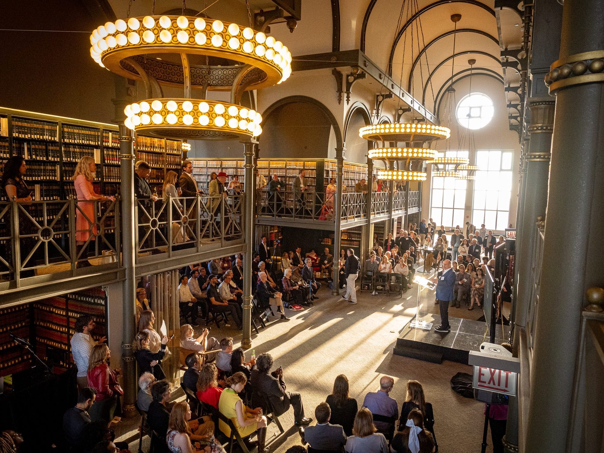 Guests seated and standing on two levels of a bookshelf-lined hall with ring chandeliers watch a man speaking below.