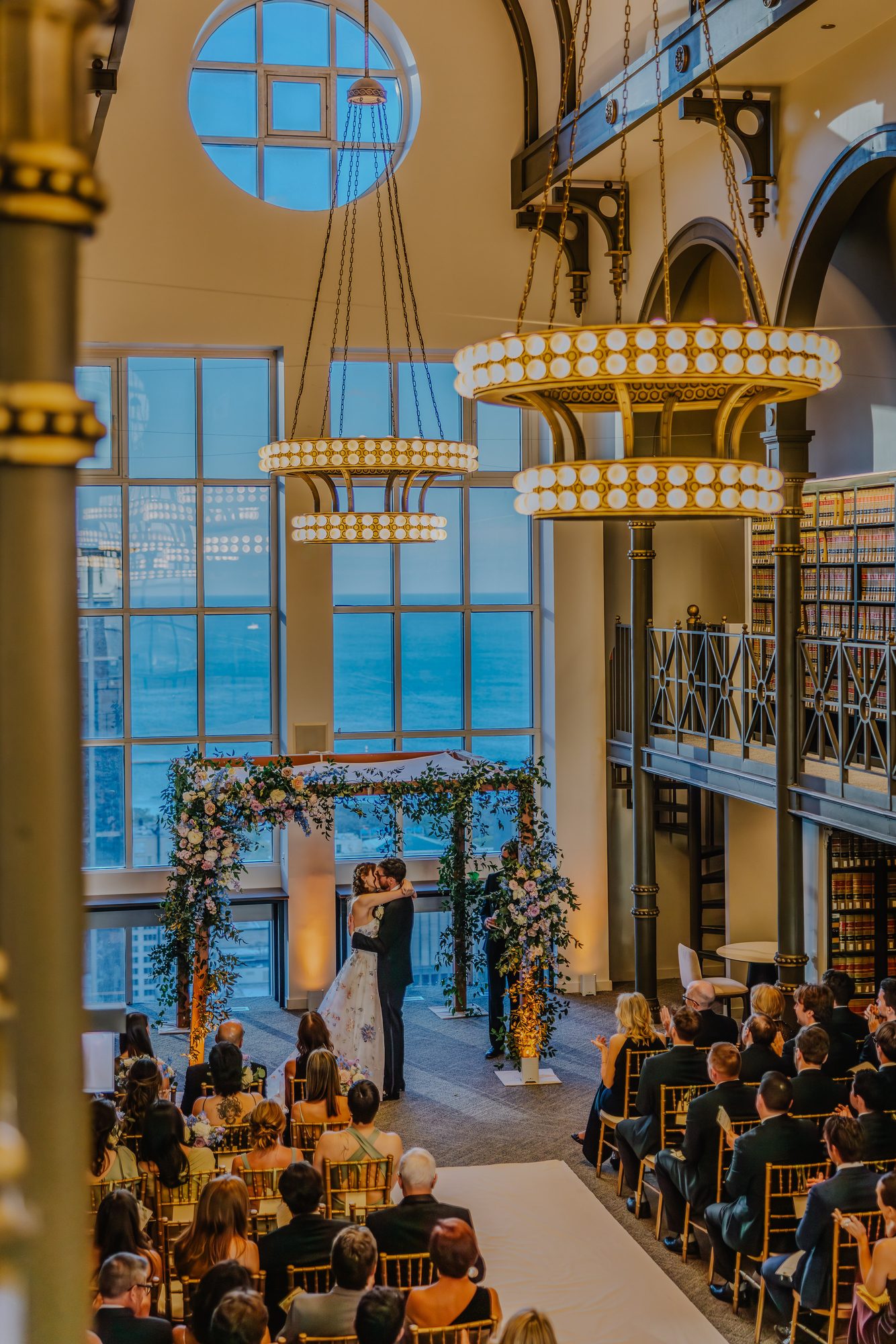 Couple kissing under a floral chuppah as seated guests watch, beneath round chandeliers and tall arched windows.