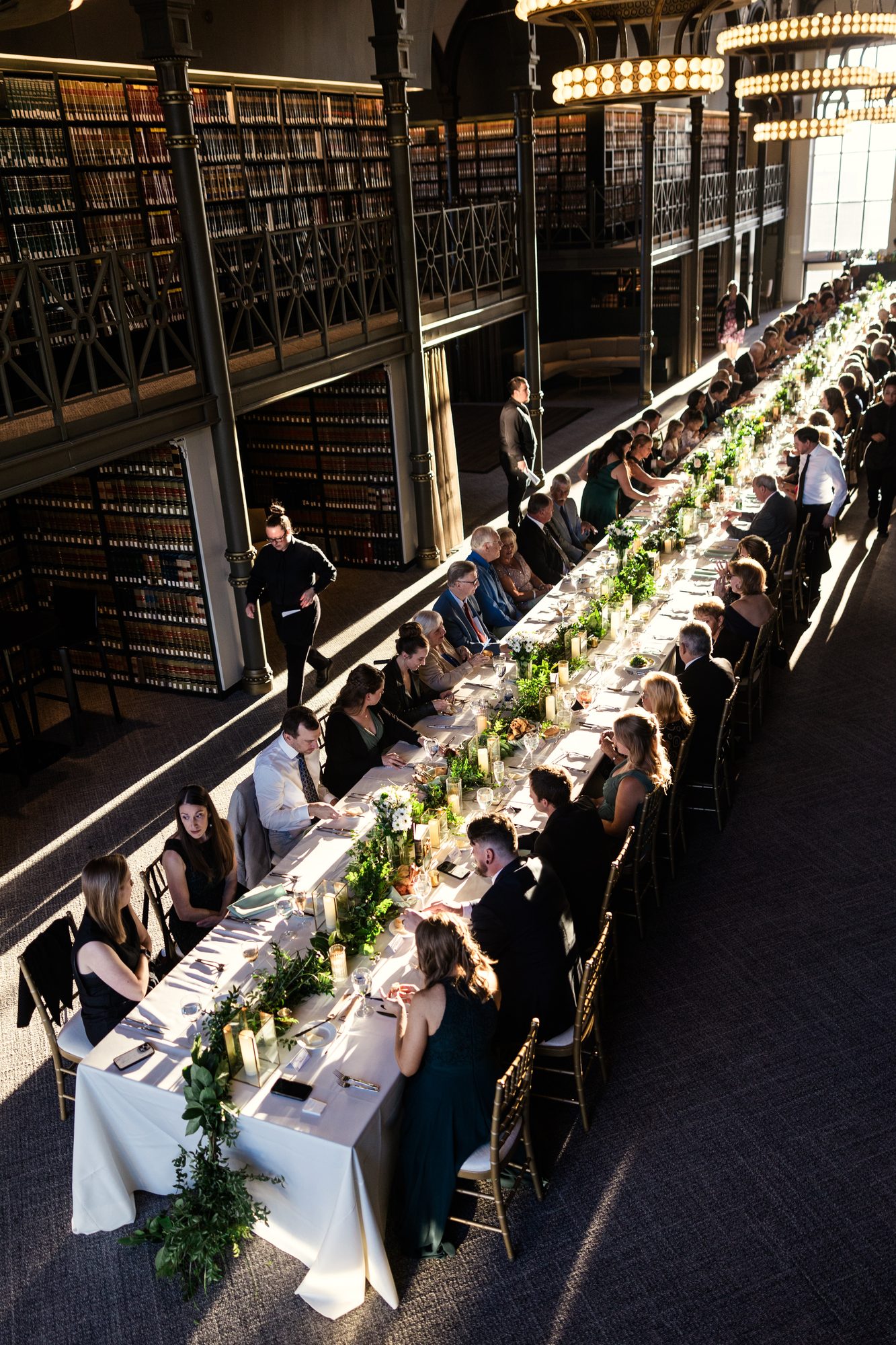 Guests in formal attire seated along one long banquet table below a book-lined mezzanine lit by chandeliers.