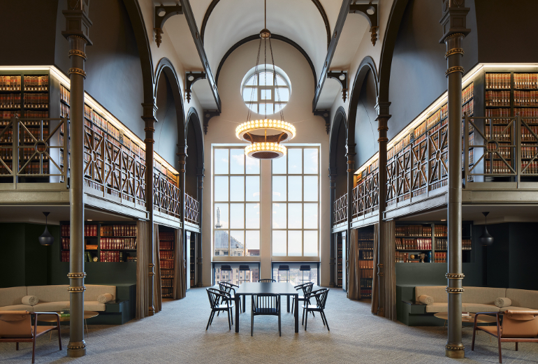 Two-story library hall with bookshelf-lined balconies, a globe chandelier, and a black table and chairs by a tall window.