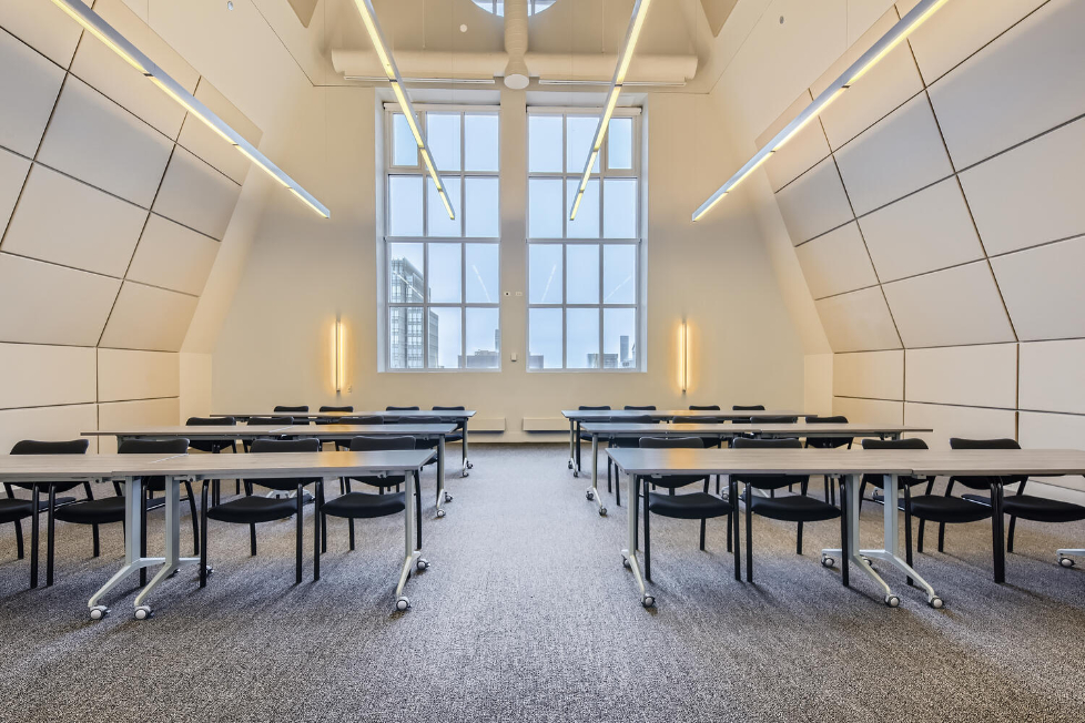 Meeting room with rows of tables and black chairs facing a tall grid-paned window under sloped white walls.