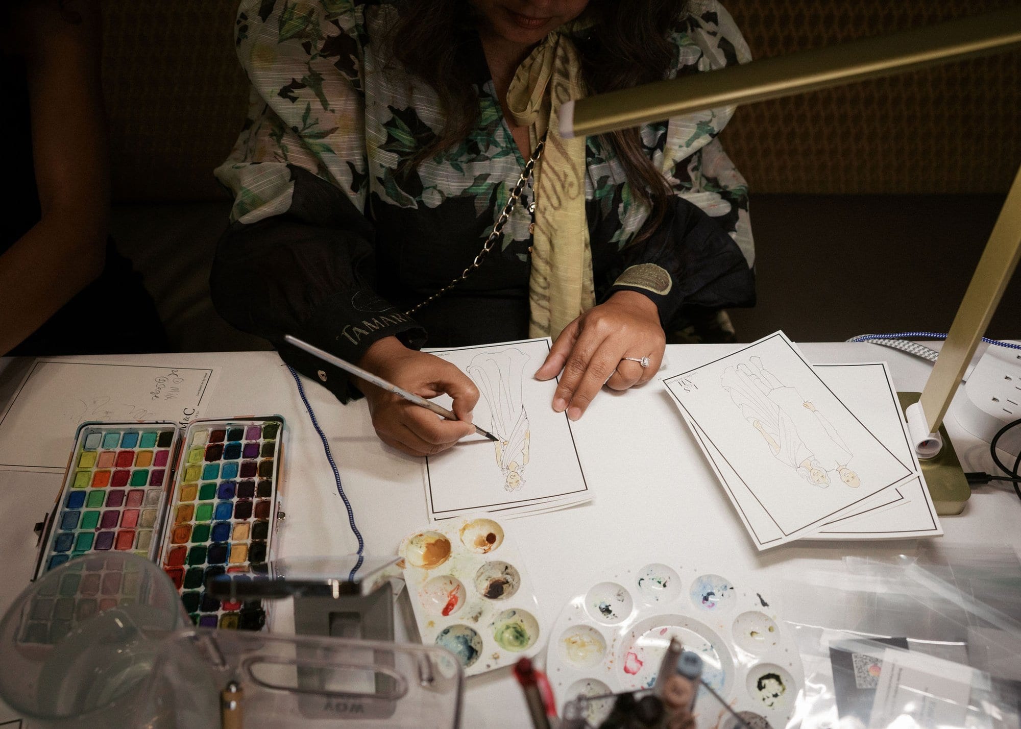 An artist sketching and painting watercolor keepsakes at a table of paints and brushes