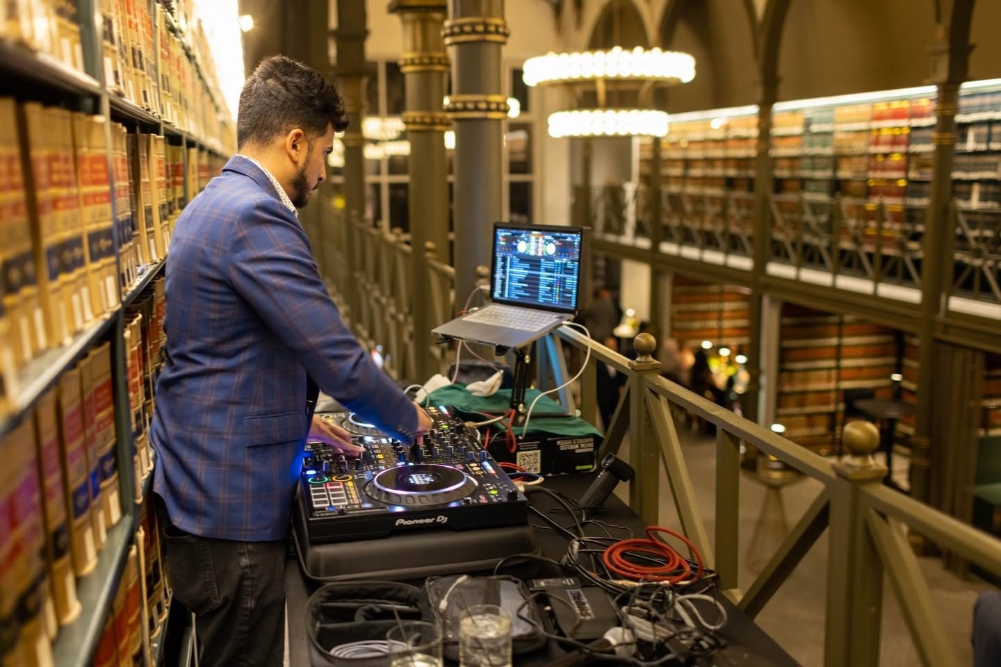 A DJ at his turntables on the balcony, with rows of bookshelves and chandeliers behind him