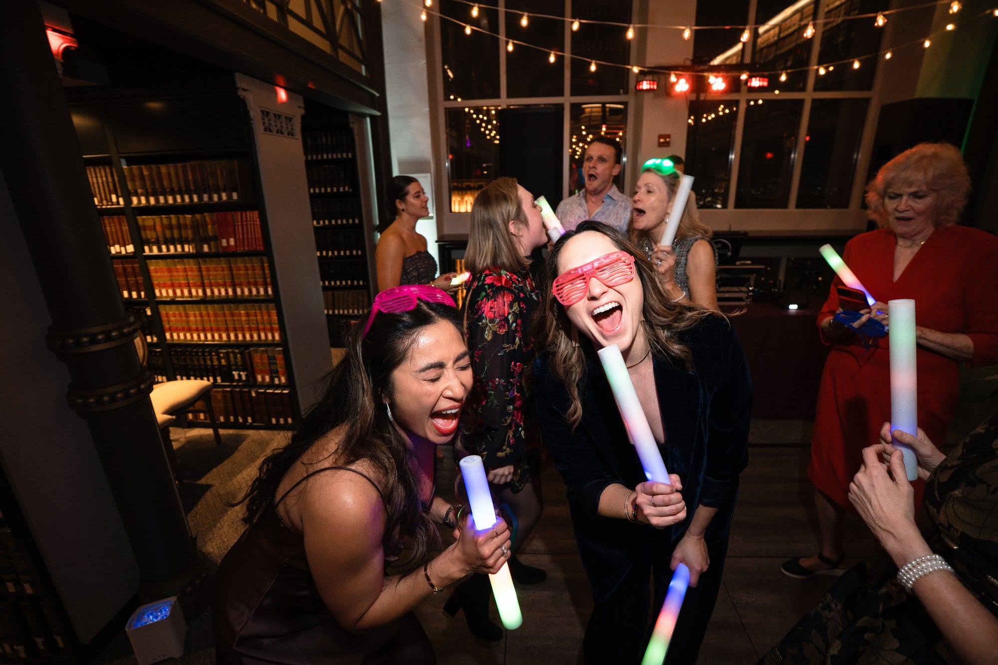Guests laughing and dancing with light-up glow sticks beneath strings of lights
