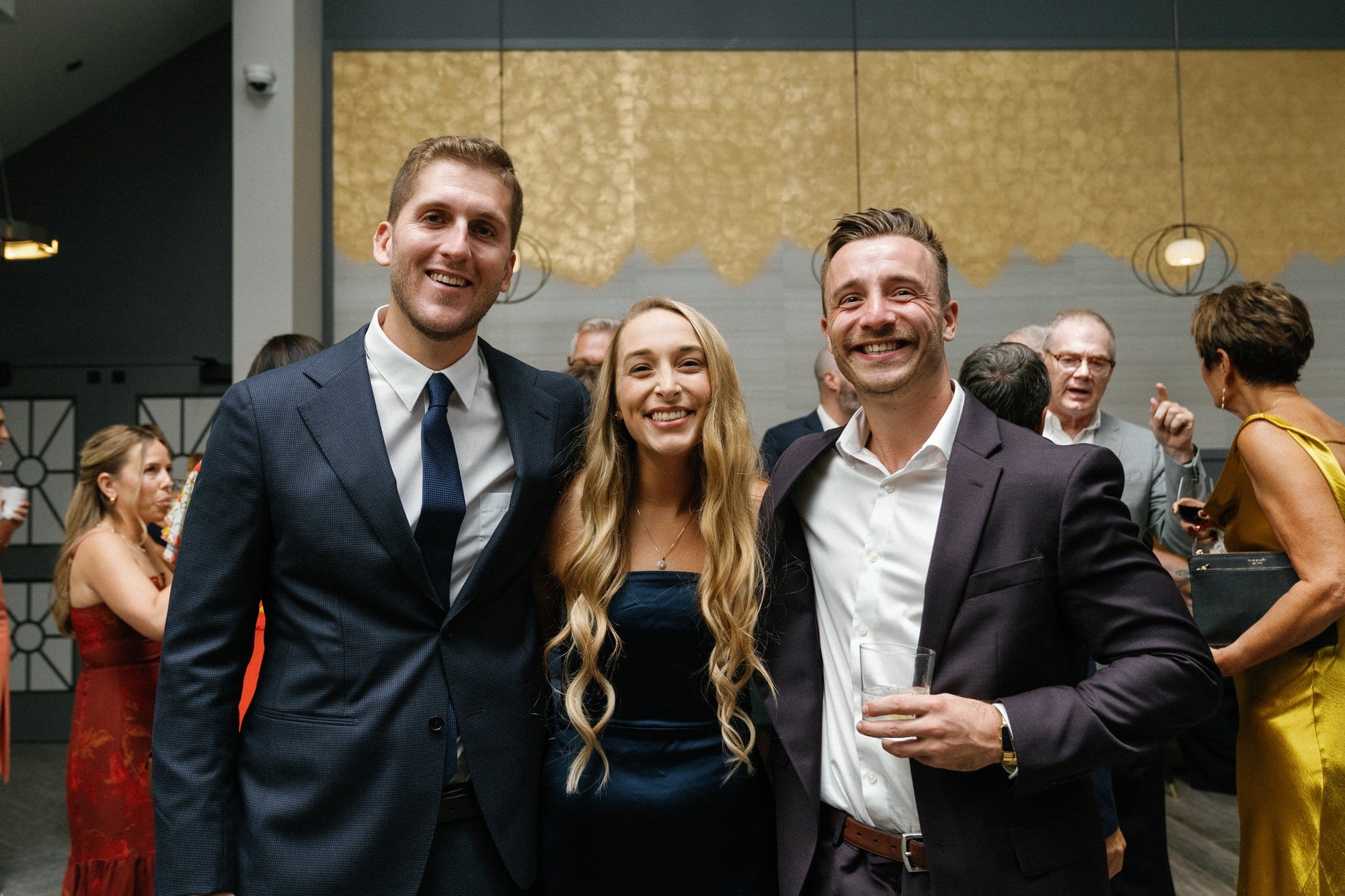 Three friends in evening wear smiling together as the party carries on around them