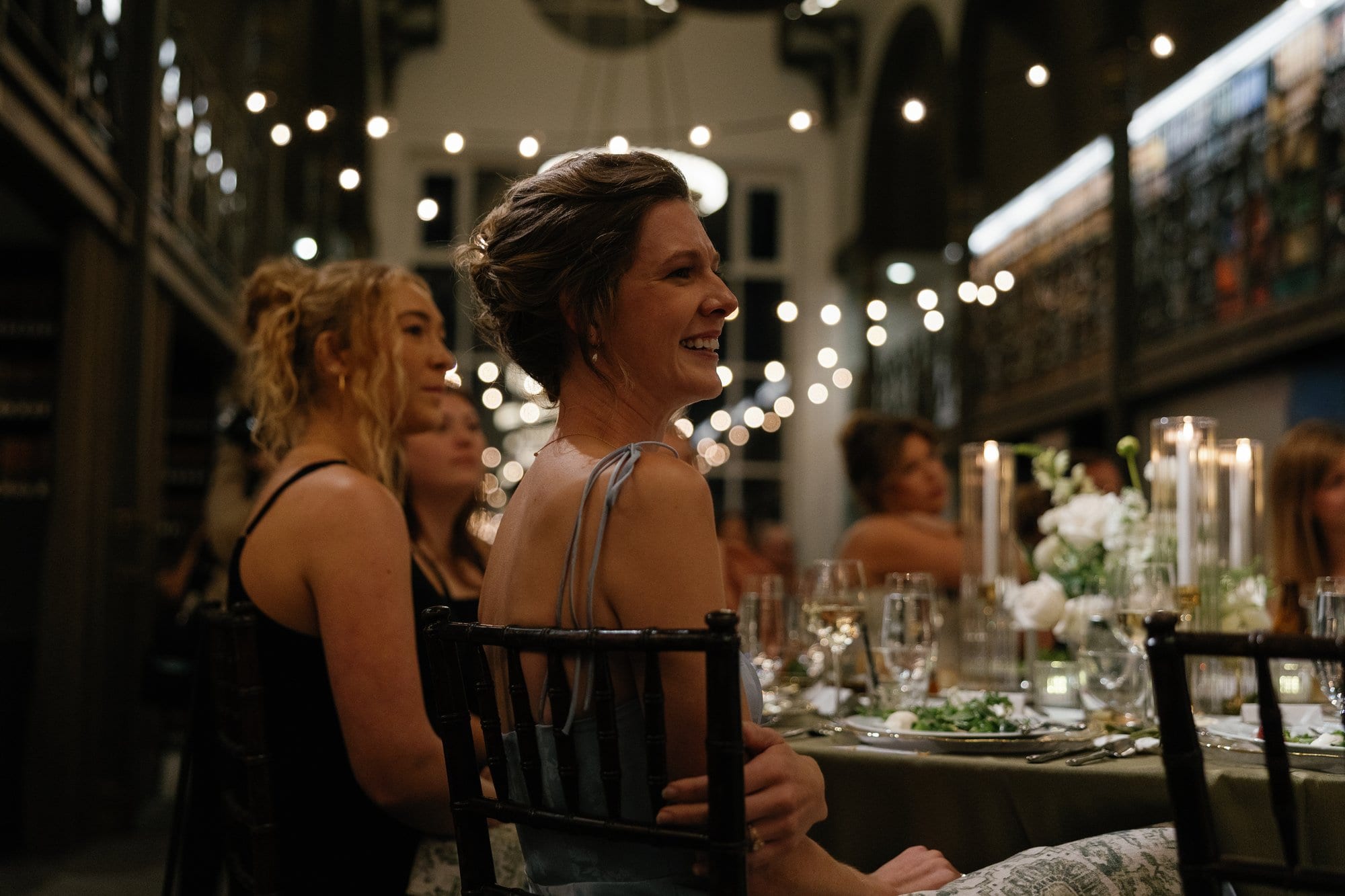 Guests smiling around a candlelit dinner table beneath strings of lights