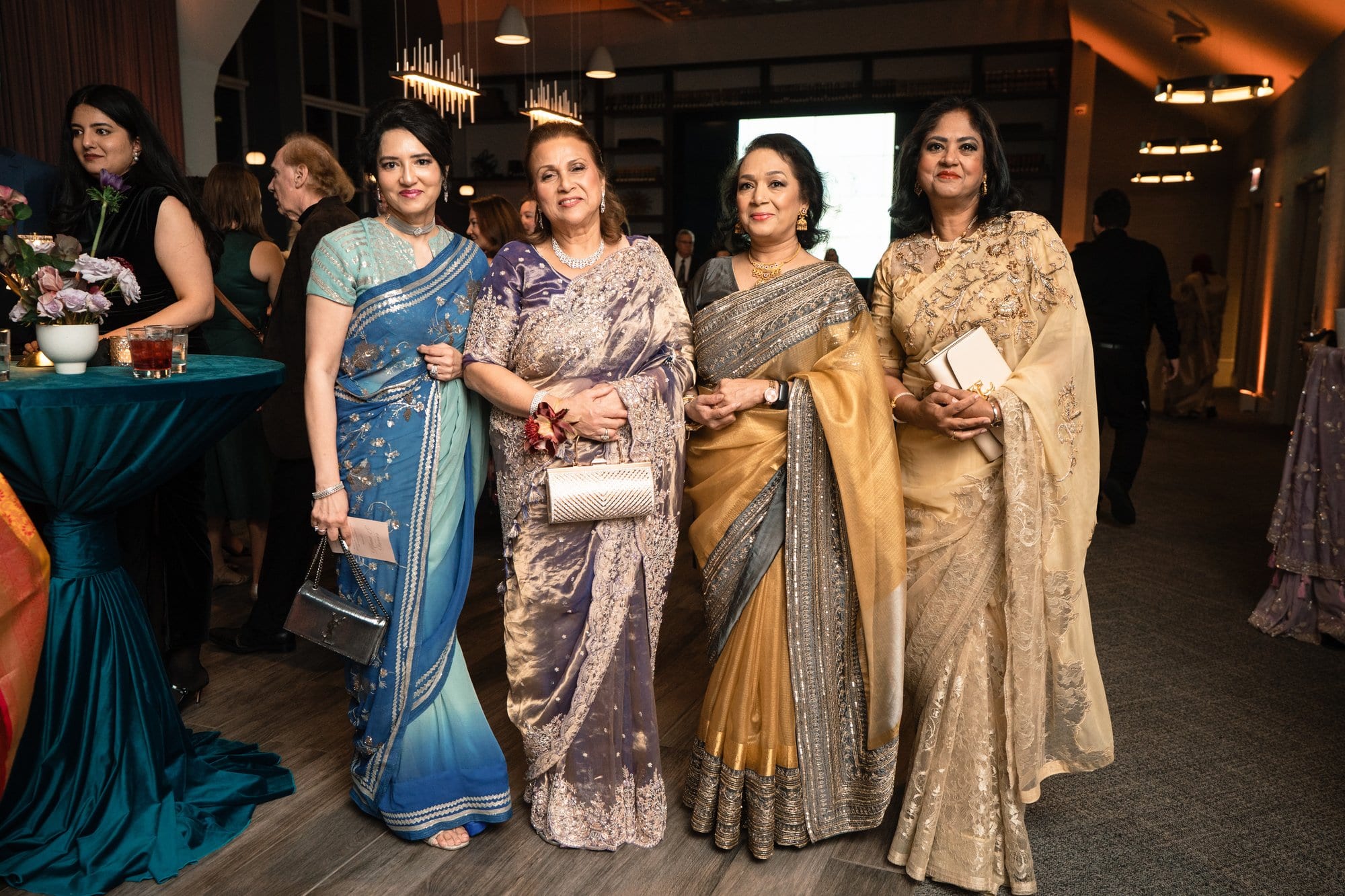Four women in shimmering saris posing together at an evening celebration